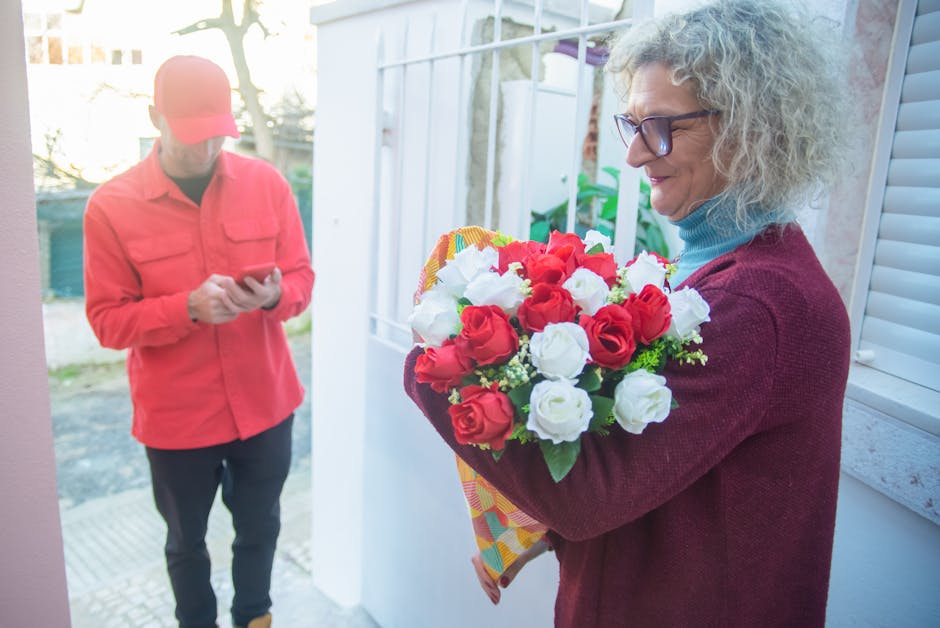 A woman with curly gray hair wearing glasses, a blue turtleneck, and a maroon cardigan holding a vibrant bouquet of fresh red and white roses wrapped in colorful paper, standing near a window in a bri