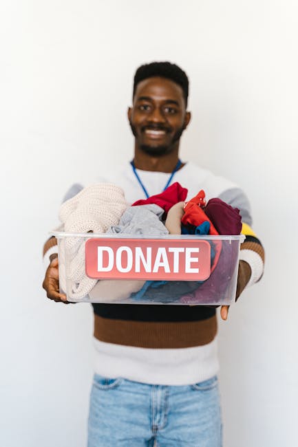 A cheerful man with dark skin and a wide smile, holding a transparent plastic box filled with a variety of colorful clothes and textiles, including beige and red fabric. The box features a prominent r