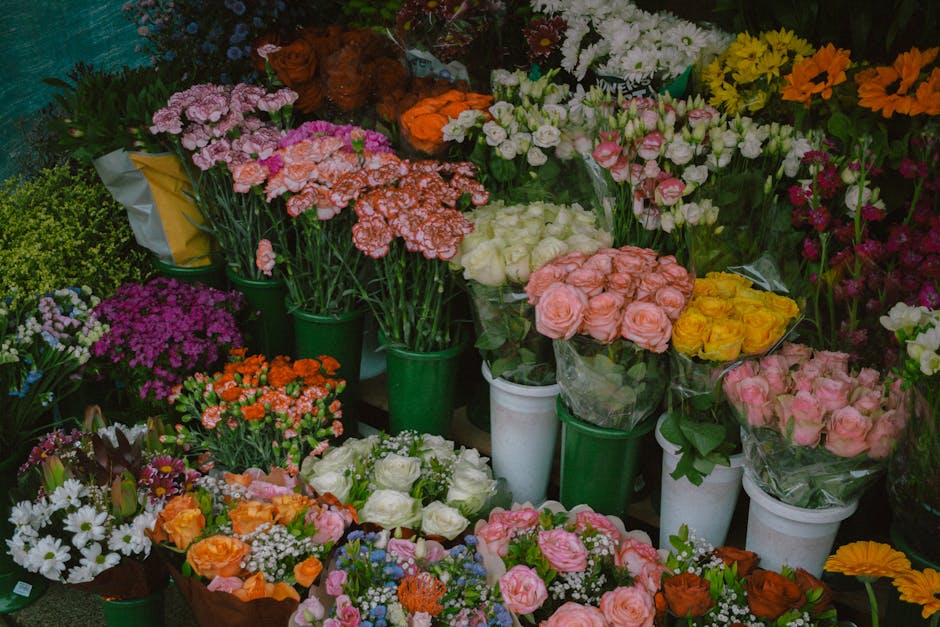 A vibrant display of fresh flowers arranged in various bouquets and potted plants, featuring a mix of roses, carnations, daisies, and other blooms in shades of pink, white, yellow, orange, and purple.