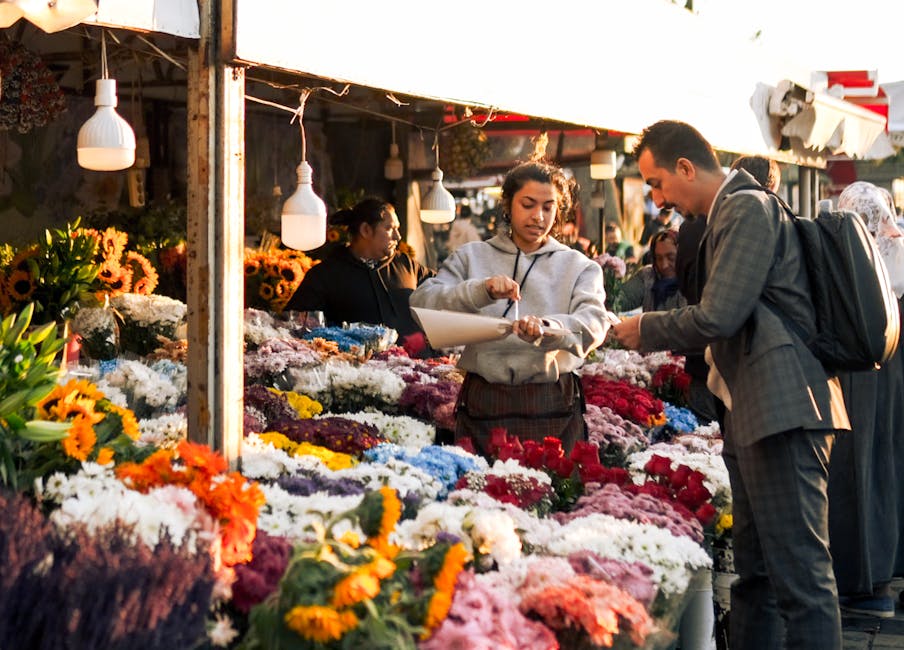 At a bustling flower market, a female florist from flowerskingstonuponthames.co.uk is engaging with a male customer over a vibrant display of fresh flowers arranged on a wooden stand. The florist, dre