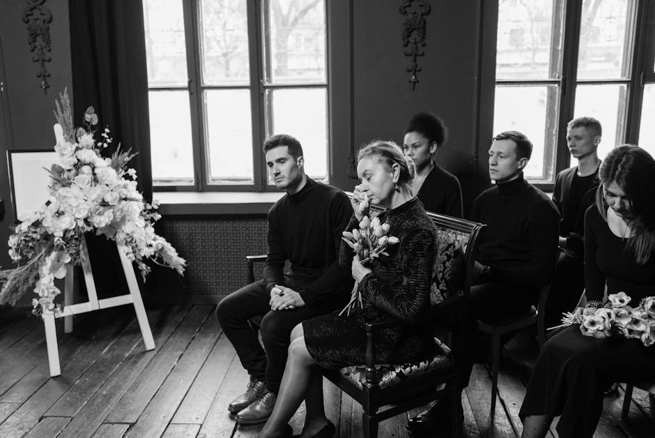 A group of six individuals sitting in a solemn setting with large windows in the background, attending a funeral or memorial service. In the foreground, a woman holds a bouquet of fresh tulips and ros