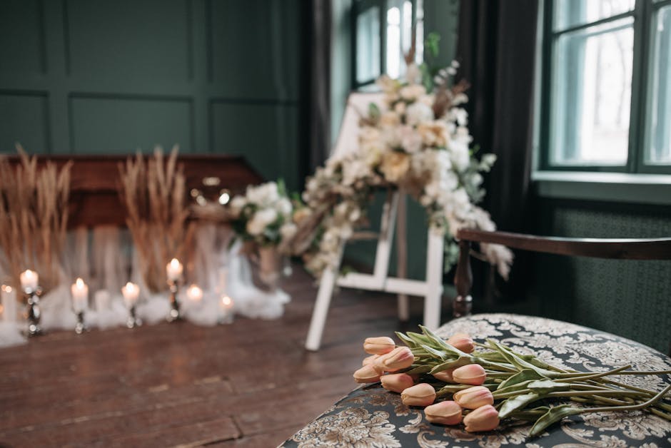 A floral arrangement setup with a bouquet of pale pink tulips resting on an ornate upholstered chair in the foreground. In the background, an elegant funeral tribute composed of white roses, lilies, a